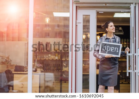 Mixed race woman worker or store owner with standing in the doorway of her grocery shop looking aside smiling. Portrait of girl assistant wearing apron welcoming inviting open door of store.