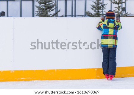 Winter sunny day. Little boy in the red skates stands on the ice, resting between laps, holding for the side of the ice rink. Training. Winter entertainment and pastime. Leisure and lifestyle.