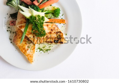 A delicious grilled chicken fillet served on a white plate with cooked broccoli and carrots aside. On a white background.