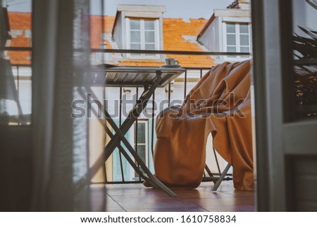 cozy terrace or balcony with small table, chair with blanket, newspaper, phone, tablet, small cup of coffee on the table. Yellow buildigs with big french windows and red roof on background.