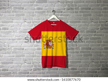 Spain flag on shirt and hanging on the wall with brick pattern wallpaper, a horizontal of red yellow and red; charged with the Spanish coat of arms left of center.
