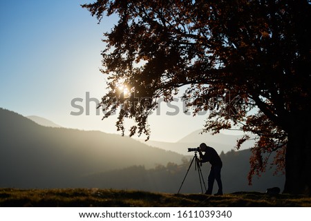 Silhouette of male tourist photographer using tripod and professional camera to take picture of mountain panorama, standing under large tree on woody foggy mountains landscape and blue sky background.