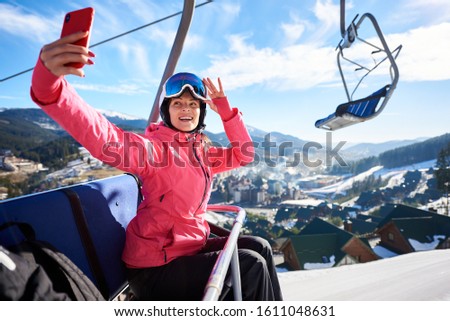 Young attractive woman, smiling skier in helmet, goggles and warm clothing riding in cable car high in air holding smartphone, taking selfie on winter ski resort background. Active lifestyle concept.