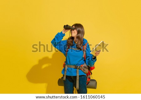 Looking forward. Portrait of a cheerful young caucasian tourist girl with bag and binoculars isolated on yellow studio background. Preparing for traveling. Resort, human emotions, vacation. Royalty-Free Stock Photo #1611060145