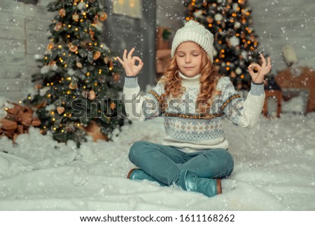 A cute blond-haired girl in knitted hats sits in the snow in winter in Turkish with closed eyes behind her is a wooden house and Christmas trees.