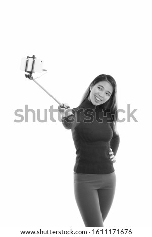 Studio shot of young happy Asian woman smiling while taking selfie picture with mobile phone while holding selfie stick