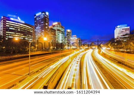 WIde multi lane Warringah freeway at the entrance to the Sydney Harbour tunnel and Bridge at sunset in front of high-rise office towers in North SYdney with bright illumination and blurred lights.