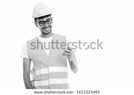 Studio shot of young happy Persian man construction worker smiling while using mobile phone