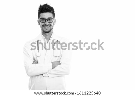 Studio shot of young happy Persian man doctor smiling while wearing eyeglasses with arms crossed