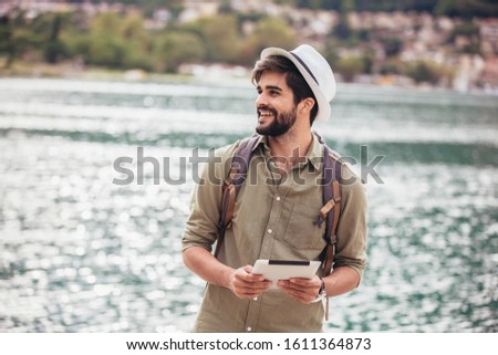 Young man walking by the harbor of a touristic sea resort with boats on background, holding digital tablet.