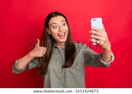 Photo of youngster lady holding telephone making selfies raising thumb finger expressing agreement talking skype friends wear casual grey green shirt isolated red color background