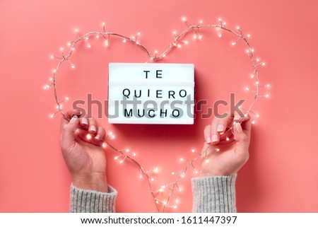 Valentine flat lay, top view on pink background. Lightbox with text "Te quiero mucho" means "I love you" in Spanish. Light garland in heart shape held in female hands. St. Valentine's day concept.