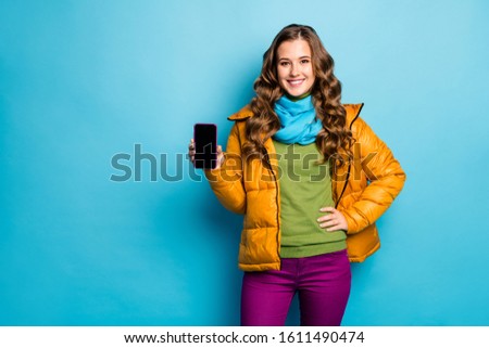 Photo of pretty lady hold telephone hands advising new gadget smart phone model low sale price wear yellow overcoat scarf violet trousers green jumper isolated blue color background