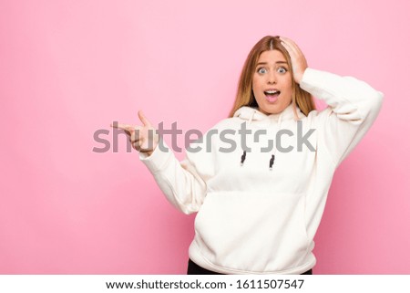 young blonde woman laughing, looking happy, positive and surprised, realizing a great idea pointing to lateral copy space against flat wall