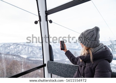 A young beautiful blonde girl in winter clothes and a gray knitted hat sits in the gondola lift cabin in the mining resort and takes photos of the mountains and landscape Altai.