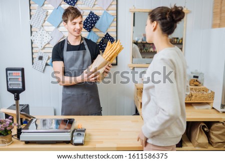 Young man bakery shop owner standing next to counter selling products in zero waste shop for beautiful female customer in grocery store. Small business,conscious minimalism vegan lifestyle concept.