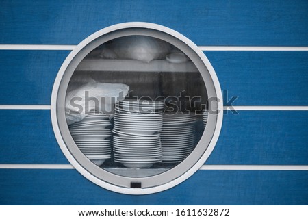 A large round porthole in the blue side of the ship, through which you can see stacked plates and dishes. restaurant kitchen on the ship, fast food Royalty-Free Stock Photo #1611632872