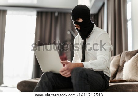 A male hacker in a mask and shirt is working on a laptop, trying to hack the company's management system