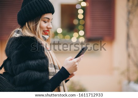 Pretty girl walking against a wall with Blurred Christmas Lights on background, she has bright makeup and red lipstick. Woman using cell phone in cold weather outside in city urban setting.
