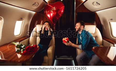 Love in the air. A beautiful couple is celebrating St Valentine’s Day on a private jet. A man is giving a present in a red box to his girlfriend, who is sitting with her eyes closed. Royalty-Free Stock Photo #1611726205
