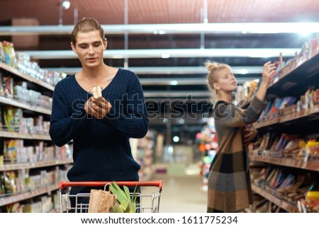 Young couple at the supermarket doing daily shopping walking with cart boyfriend reading ingredients curious while girlfriend choosing chocolate excited Royalty-Free Stock Photo #1611775234
