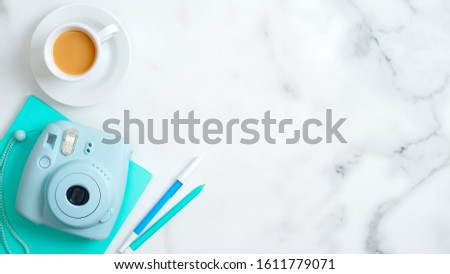 Trendy modern instant camera, cup of coffee, mint paper notebook with office supplies on marble desk. Flat lay, top view, copy space. Stylish feminine home office desk table with elegant accessories.