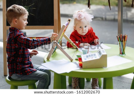 Children draw. Blonde girl sits at a table, a boy stands near a blackboard.