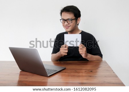 Young Asian man feeling happy with laptop and holding empty white space paper. Smile Indonesia Man wear black shirt Isolated grey background.