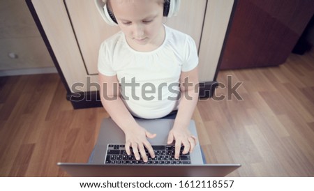 A smart little girl of seven years old in white headphones with a laptop in her hands is pushing on the floor in her room. The young generation on the Internet and IT technology