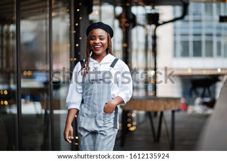 African american woman in overalls and beret posed in outdoor terrace with christmas decorations garland.