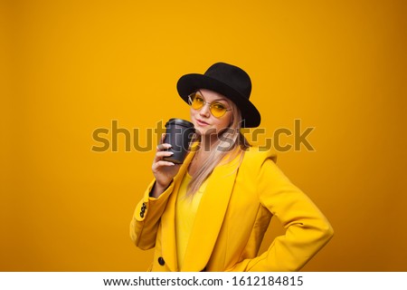 Take-away coffee. Stylish trendy young woman in bright clothes on a yellow background. A cool blonde girl in a yellow jacket, yellow sunglasses and a black hat drinks coffee and is happy