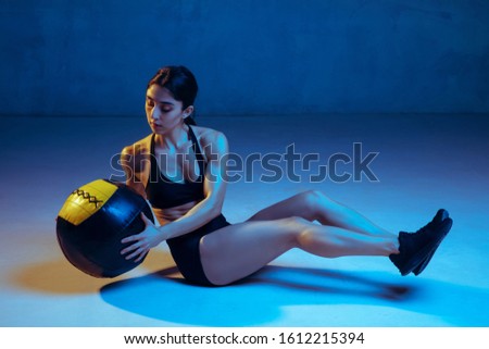 Caucasian young female athlete practicing on blue studio background in neon light. Sportive model training her lower body with ball. Body building, healthy lifestyle, beauty and action concept.