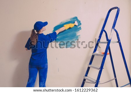 A young woman in a blue T-shirt in yellow rubber gloves and cap is painting a wall in an apartment, there is a staircase nearby.