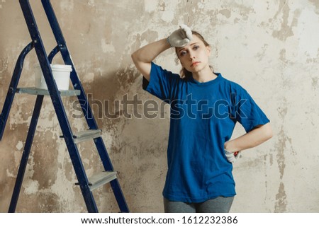 A young woman in work clothes wearily wipes sweat from her forehead after preparing the plastered wall for painting.
