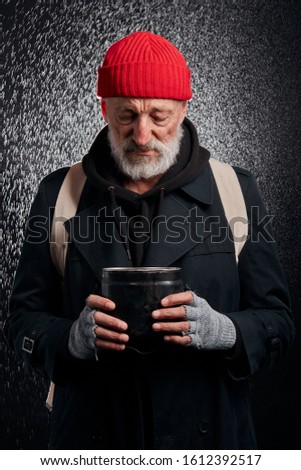 Bearded old man wearing black street clothes and holding iron cup for money, for help. Sad homeless man under heavy rain.