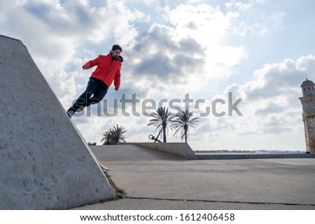 Young man in a red jacket ready to jump off a side wall in a square with an antique lighthouse