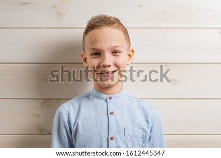 A boy of 10 years old in a blue shirt smiles on a light wooden background and makes various signs with his hands.