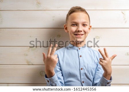 A boy of 10 years old in a blue shirt smiles on a light wooden background and makes various signs with his hands.