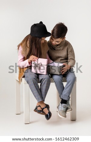 Cute stylish little couple girl and boy in fashionable clothes sittting together at studio. child boy gives smiling girl pink gift box. St. Valentine's Day