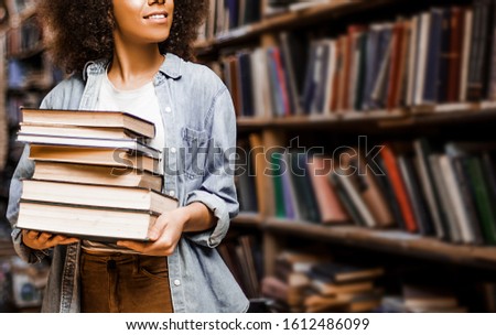 Photo for background, place for insertion, African American woman student holding a heavy armful of books in her hands against the background of bookshelves in a library. Learning concept, lifestyle