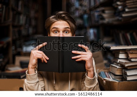 Stunned guy hiding behind a book, peeps out with surprised shocked eyes. New interesting information, Education lover surrounded by books in the library. student lifestyle. Reading time.  Royalty-Free Stock Photo #1612486114