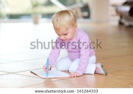 Adorable lovely baby girl plays indoors drawing with colorful pencils sitting on tiles floor