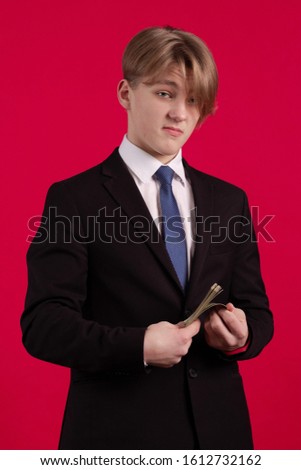 Teen boy in a black jacket holds a bundle of dollars banknotes in his hands and poses on a red background