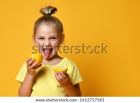 Little smiling cute blond girl in yellow t-shirt holding halves of fresh sour ripe lemon fruit and showing tongue over yellow background. Healthy lifestyle and clean eating concept