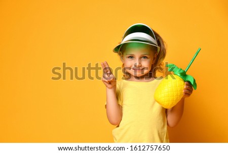 Little smiling cute blond girl in yellow t-shirt and hat holding fresh healthy fruit juice in pineapple shaped bottle with straw over yellow background. Healthy lifestyle and clean eating concept