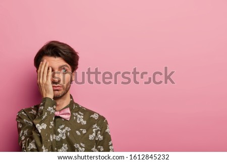 Studio shot of serious youngster with calm expression, covers face with palm, coves half of face with palm, dressed in stylish outfit, isolated over pink background, empty space area for information