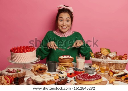 Cheerful mixed race woman eats with big appetite creamy pancakes, has unhealthy diet, looks gladfully, being sweet tooth, overeats desserts, isolated over pink background, cannot prevent obesity Royalty-Free Stock Photo #1612845292