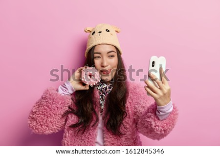 Dark haired teenager licks lips, poses with delicious sweet glazed donut, returns from bakery store, takes selfie on mobile phone, has fun, wears winter outft, models over pastel rosy background