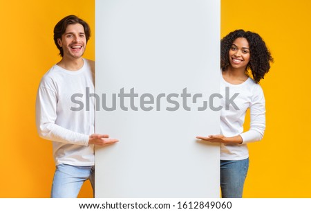 Cheerful Guy And Afro Girl Holding And Pointing At White Advertisement Board, Standing Together Over Yellow Background Royalty-Free Stock Photo #1612849060