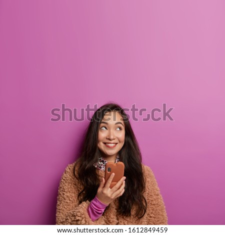 People and technologies concept. Dreamy lovely dark haired girl keeps mobile phone in hand, sends voice message to friend, concentrated above with smile, waits for call, blank space upwards.